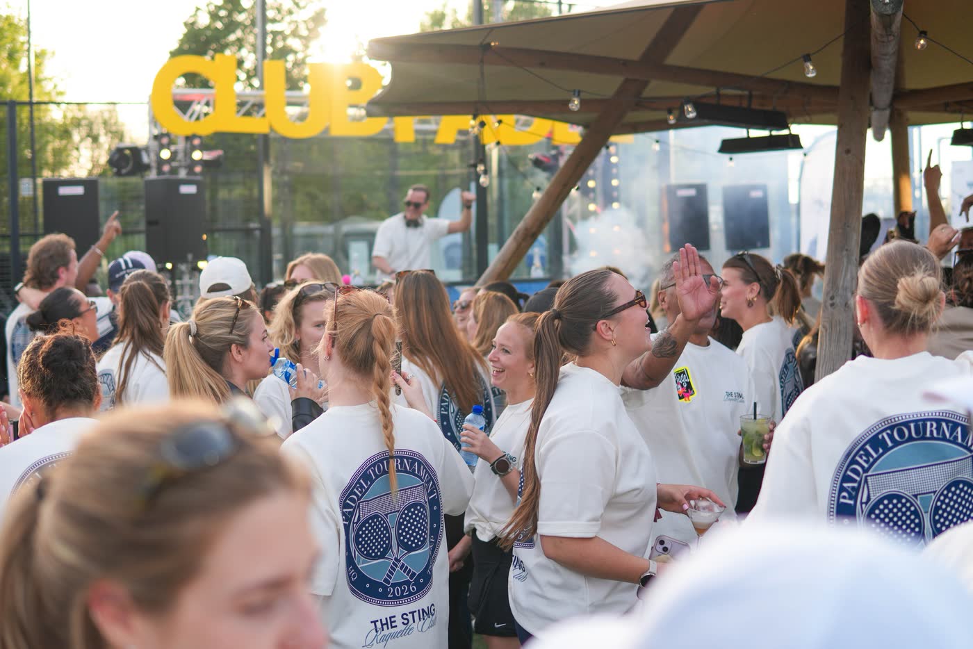 Deelnemers in witte toernooishirts feesten onder een overkapping, met gele Club Padel-letters op de achtergrond
